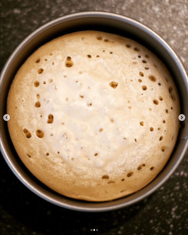 A freshly baked round loaf of bread with a golden crust and numerous small holes, known as air pockets, sits inside a round baking tin. The bread has a fluffy texture and is viewed from the top down, resting on a speckled dark countertop background.